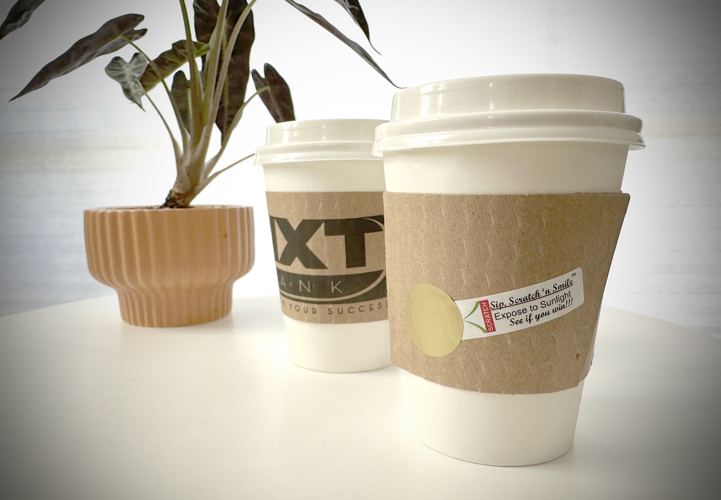Custom Cup Sleeves with scratch off feature applied to kraft cup sleeves placed on 2 white paper cups placed on a tabletop with plant in background