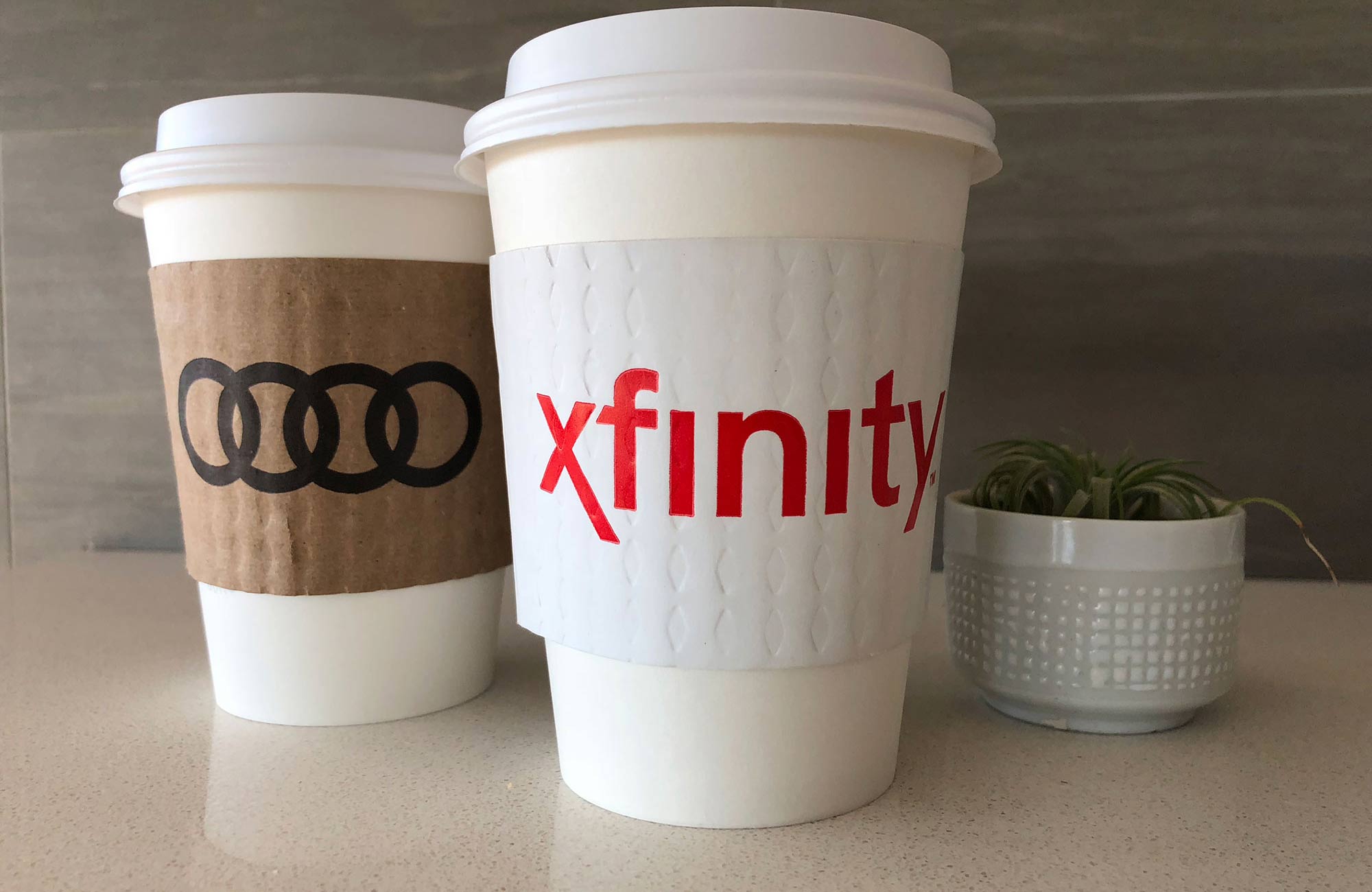 Custom cup sleeves on printed in 1 color ink on white and kraft sleeves for Xfinity and Audi and are placed on white paper cups displayed on countertop with plant in backround