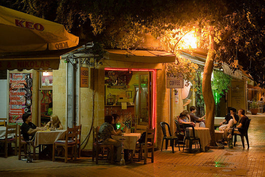 A nighttime street scene outside a small cafe with outdoor seating, warm yellow lighting, people dining at tables, and a tree in the foreground on a paved walkway.