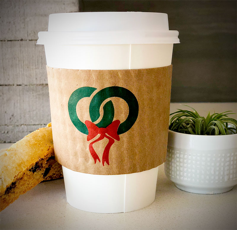 Kraft custom printed coffee cup sleeves printed with two colors on front and back placed on a white paper cup placed on tabletop with plants on background