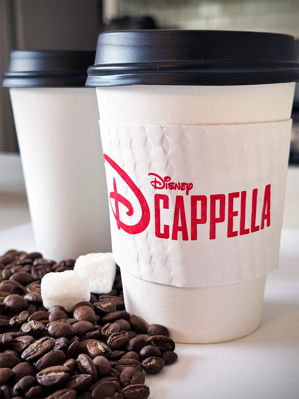 Printed coffee cup sleeves printed on white sleeves with one color on front placed on 2 white paper cups on a table with coffee scattered coffee beans